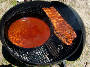 Weber grill set up for indirect heating, with charcoal only on one side separated by bricks. Ribs on indirect, with sauce directly over the fire to simmer.